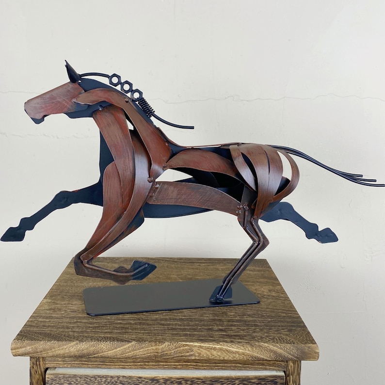 May include: A metal sculpture of a running horse. The horse is crafted from brown and black metal pieces, with a flowing mane and tail. The sculpture is mounted on a black base and sits on a wooden surface.