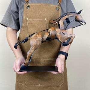May include: A brown metal sculpture of a horse in mid-gallop. The sculpture has a distressed finish, with black accents and a black base. The horse's mane and tail are detailed, and the sculpture is held by a person.