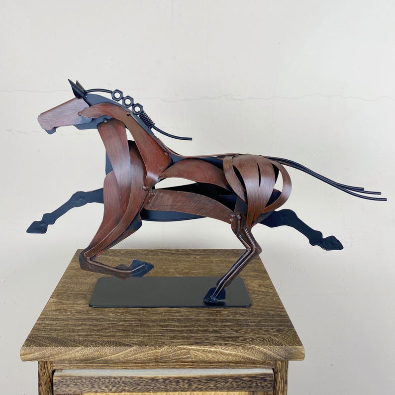 May include: A metal sculpture of a running horse. The horse is crafted from brown and black metal pieces, with a flowing mane and tail. The sculpture is mounted on a black base and sits on a wooden surface.