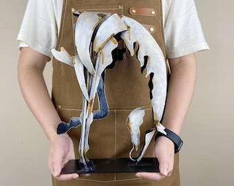 May include: Abstract metal sculpture with white, black, and gold accents. The artwork features a curved, flowing design mounted on a black rectangular base. The sculpture is held in front of a person wearing a brown apron.