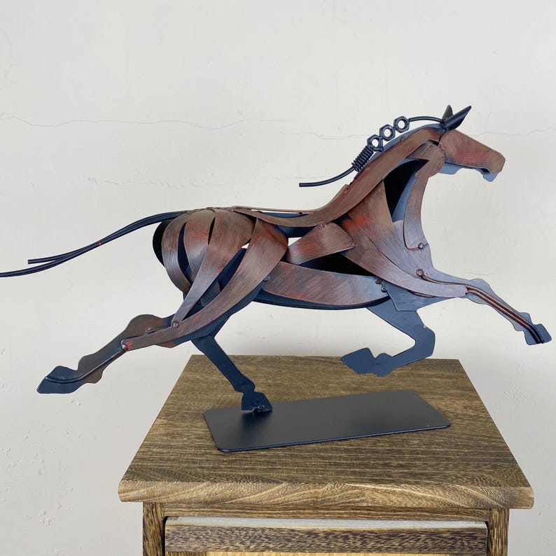 May include: A metal sculpture of a running horse. The sculpture is made of layered metal pieces in shades of brown, red, and black. The horse is mounted on a black rectangular base and is displayed on a wooden table.
