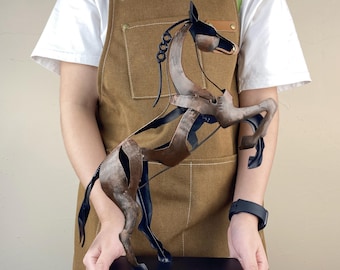 May include: A metal sculpture of a rearing horse. The sculpture is primarily brown with black accents, and it is mounted on a black rectangular base. The horse's mane and tail are detailed with curved metal pieces. The sculpture is held in front of a person wearing a brown apron.