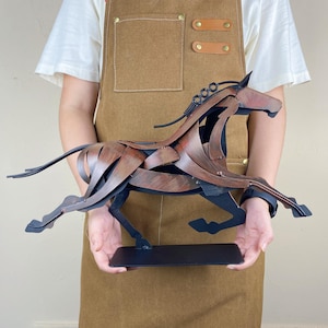 May include: A metal sculpture of a running horse. The horse is made of layered, textured metal in shades of brown and black. The sculpture is mounted on a rectangular black base. The horse's mane is made of coiled metal.