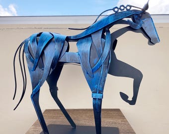 May include: A blue metal horse sculpture with a stylized design. The sculpture features a dark blue base and a lighter blue body with intricate cutouts. The horse is posed in a dynamic stance, with its front leg raised. The sculpture is displayed on a wooden surface.