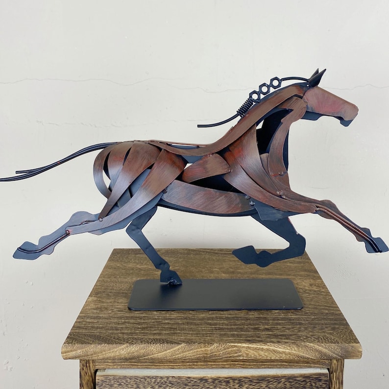 May include: A metal sculpture of a running horse. The horse is constructed from layered metal pieces in shades of brown and gray, with a black base. The sculpture is mounted on a rectangular black base and is displayed on a wooden surface.