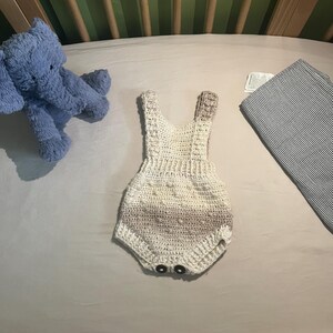 May include: A crocheted baby onesie in white and brown with a gradient pattern. The onesie has a sleeveless design and two black buttons at the bottom.