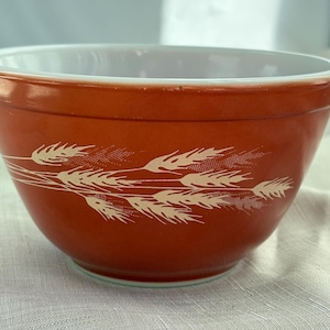 May include: A vintage orange mixing bowl with a white interior and a wheat stalk design. The bowl has a rounded shape and is made of a durable material, perfect for kitchen use. The wheat design adds a touch of rustic charm.