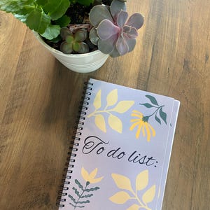 Peut inclure: Un carnet à spirales avec une couverture violet clair ornée de l'inscription "To do list:" en écriture cursive. La couverture est décorée de motifs floraux jaunes et verts. Un petit pot blanc avec des succulentes vertes et violettes est à côté.