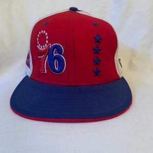 Könnte beinhalten: Rote, weiße und blaue Baseballkappe mit der gestickten Zahl "76" auf der Vorderseite. Die Kappe hat einen blauen Schirm und blaue Sterne an der Seite. Ein sportliches Accessoire.