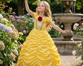 Può includere: Un abito giallo di raso con gonna a balze e maniche corte. L'abito presenta un dettaglio di rosa rossa sullo scollo e finiture dorate. La ragazza è in un giardino con una fontana sullo sfondo.