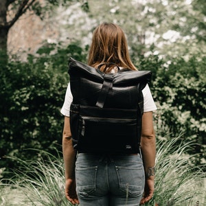 Pode incluir: Uma pessoa vestindo uma camisa branca e jeans azul está carregando uma mochila preta de rolagem com um bolso com zíper e um pequeno logotipo na frente.