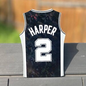 Pode incluir: Um ornamento em forma de camisola de basquetebol preta com um padr&atilde;o geom&eacute;trico hologr&aacute;fico. O nome HARPER est&aacute; impresso em letras mai&uacute;sculas brancas acima do n&uacute;mero 2, que tamb&eacute;m &eacute; branco com um contorno preto. As bordas t&ecirc;m um acabamento branco.