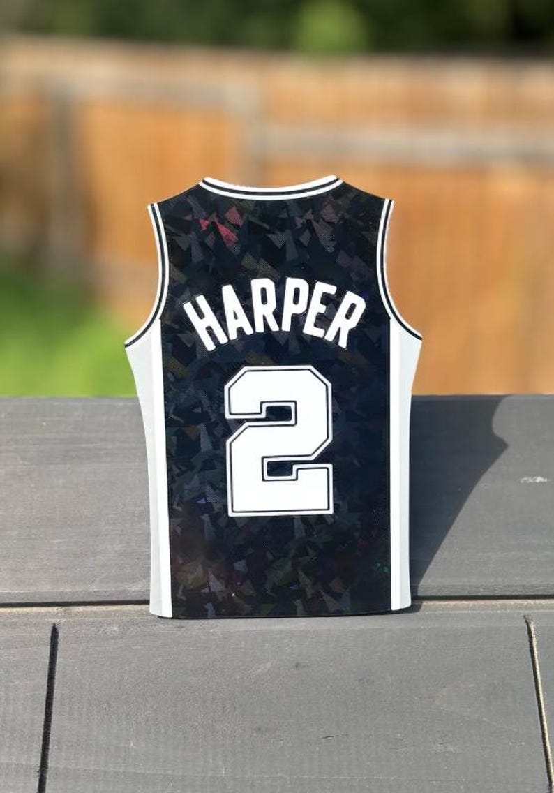 Pode incluir: Um ornamento em forma de camisola de basquetebol preta com um padr&atilde;o geom&eacute;trico hologr&aacute;fico. O nome HARPER est&aacute; impresso em letras mai&uacute;sculas brancas acima do n&uacute;mero 2, que tamb&eacute;m &eacute; branco com um contorno preto. As bordas t&ecirc;m um acabamento branco.