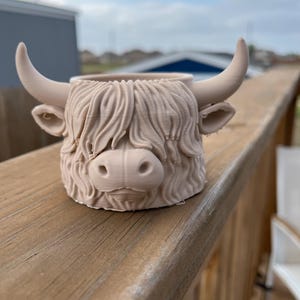 May include: A beige-colored Highland cow planter with detailed fur, horns, and facial features. The planter is designed to hold small plants and adds a unique touch to any home decor. The planter is made of a durable material.