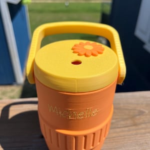 May include: An orange and yellow water cooler with a handle. The cooler has the name "Michelle" engraved on the side. The lid is yellow with an orange flower and a small hole. A yellow spigot is at the bottom.