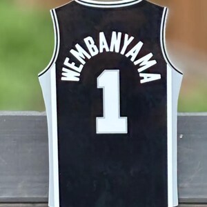 Pode incluir: Uma camisola de basquetebol preta com acabamento branco ao longo do decote e das laterais. A parte de tr&aacute;s apresenta o nome WEMBANYAMA em letras brancas arqueadas sobre o n&uacute;mero 1, tamb&eacute;m em branco com um contorno cinzento no tecido.
