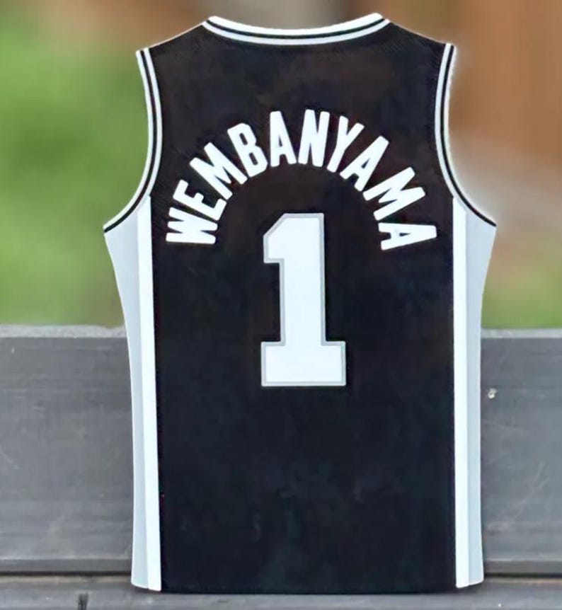 Pode incluir: Uma camisola de basquetebol preta com acabamento branco ao longo do decote e das laterais. A parte de tr&aacute;s apresenta o nome WEMBANYAMA em letras brancas arqueadas sobre o n&uacute;mero 1, tamb&eacute;m em branco com um contorno cinzento no tecido.