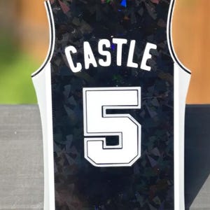Pode incluir: Uma camisola desportiva sem mangas com base preta e um padr&atilde;o refletor de estilha&ccedil;os de vidro. A parte de tr&aacute;s apresenta o nome CASTLE em letras brancas sobre o grande n&uacute;mero 5 branco, ambos com contorno preto e acabamento branco.