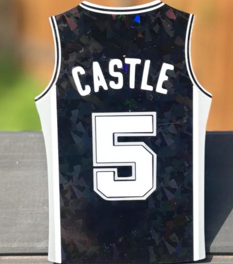 Pode incluir: Uma camisola desportiva sem mangas com base preta e um padr&atilde;o refletor de estilha&ccedil;os de vidro. A parte de tr&aacute;s apresenta o nome CASTLE em letras brancas sobre o grande n&uacute;mero 5 branco, ambos com contorno preto e acabamento branco.