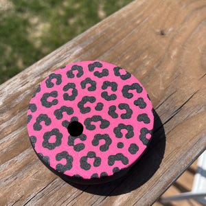 May include: A round, pink drink coaster with a black leopard print pattern. The coaster has a black border and a central hole. The coaster is sitting on a wooden surface.