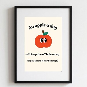May include: Framed print with the text "An apple a day will keep the a**hole away (if you throw it hard enough)". The image features a cartoon apple with a smiling face. The frame is black.