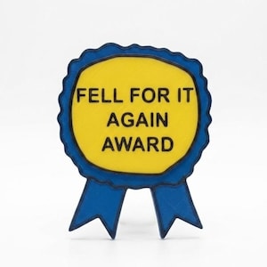May include: A blue and yellow award ribbon with the text "FELL FOR IT AGAIN AWARD". The ribbon has a scalloped edge and two blue ribbon tails. The text is in a bold, sans-serif font.