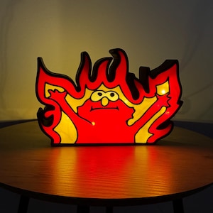 Könnte beinhalten: Eine rote und gelbe Comicfigur in Form einer Flamme, die mit einem warmen Schein beleuchtet wird. Die Figur hat große Augen und ein breites Lächeln. Die Figur ist aus Holz und ist ein dekoratives Licht.