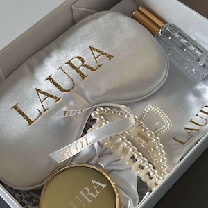 Könnte beinhalten: Eine weiße Geschenkbox mit Schlafmaske, Parfümflasche, Kompaktspiegel, Haarspange und Satinbeutel. Schlafmaske und Spiegel sind mit "LAURA" in Gold beschriftet. Ein Band trägt die Aufschrift "TO BE".