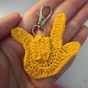 May include: A yellow crochet keychain in the shape of the American Sign Language (ASL) sign for "I love you." The keychain has a silver-colored metal clasp. The crochet work is detailed and the color is a bright, sunny yellow.