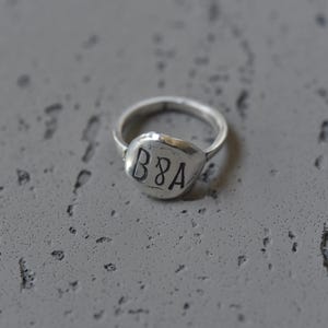 Può includere: Un anello d'argento con una faccia arrotondata e strutturata. La faccia dell'anello ha le lettere "B8A" incise. L'anello è posto su una superficie grigia e strutturata.