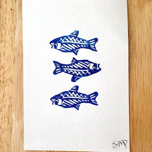May include: A white card features three blue fish prints arranged vertically. Each fish is detailed with cross-hatching and line patterns. The artist's initials, "SMP," are in the lower right corner. The card rests on a wooden surface.