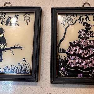May include: Two framed silhouette art pieces. One depicts two figures on a tree branch, the other a woman in a layered dress with a goose. Both have black frames and a cream-colored background, with a hanging loop.