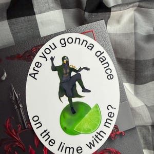 Peut inclure: Un autocollant circulaire blanc présentant l'illustration d'une silhouette vêtue d'une cape vert foncé dansant sur un citron vert. Le texte "Are you gonna dance on the lime with me?" est imprimé en noir, suivant la courbe de l'image.