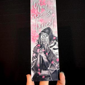 May include: A rectangular bookmark featuring a hooded figure in black and white with a mask, surrounded by falling pink cherry blossom petals. The text "You are so loved" is printed in white cursive script at the top of the bookmark.