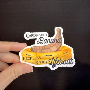 May include: A die-cut sticker featuring a brown cardboard banana sitting in a yellow inflatable lifeboat. The text reads Cardboard Banana RECKLESS on my Lifeboat in dark blue lettering against a white background with a thin white border.
