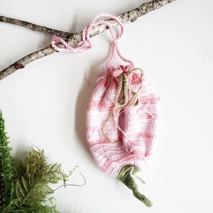 May include: A handmade, pink and white crocheted drawstring bag, resembling a flower. The bag has a green stem and is suspended from a branch with a pink cord. The bag's design features a striped pattern.