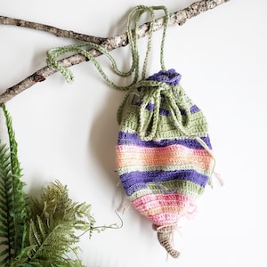 May include: A handmade crochet drawstring bag in shades of green, purple, pink, and orange. The bag is suspended from a green crocheted strap and features a decorative tassel at the bottom. The bag is hanging from a tree branch.