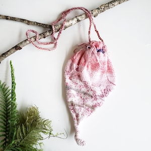May include: A handmade, pink and white crocheted drawstring bag with a long, braided strap. The bag is suspended from a natural branch against a white background. The bag's design features a pointed bottom and a gathered top, with a small loop at the end.