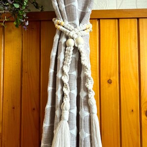 Op de afbeelding: Crèmekleurige macramé gordijn tiebacks met houten kralen en kwasten. De tiebacks houden een patroon gordijn vast. De achtergrond is een houten muur.