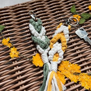 Könnte beinhalten: Zwei handgefertigte Makramee-Schlüsselanhänger in einem weißen, grünen und gelben Farbschema. Die Schlüsselanhänger hängen an einem Weidenkorb mit getrockneten Blumen.
