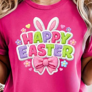 Könnte beinhalten: Ein magentafarbenes T-Shirt mit dem Schriftzug "HAPPY EASTER" in bunten, gestickten Buchstaben. Das Design umfasst eine rosa Schleife, Hasenohren und florale Akzente, perfekt für Ostern.
