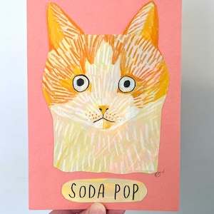 May include: A colorful cat portrait on a pink background. The cat has orange and white fur, large eyes, and a small nose. The text "SODA POP" is below the cat's image on a yellow label. The artwork is held by a hand.