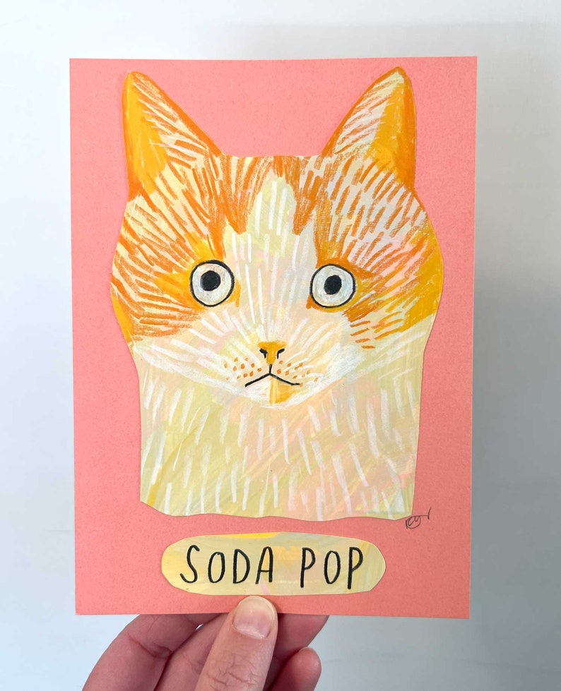 May include: A colorful cat portrait on a pink background. The cat has orange and white fur, large eyes, and a small nose. The text "SODA POP" is below the cat's image on a yellow label. The artwork is held by a hand.