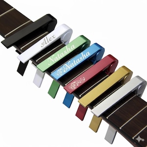 Op de afbeelding: Een verzameling gitaarcapo's in diverse kleuren: zwart, wit, groen, blauw, rood, goud en zilver. Elke capo is gepersonaliseerd met een naam of muzieknoten, rustend op een gitaar toets.