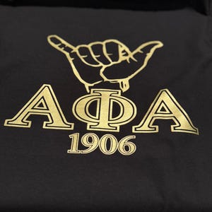 以下が含まれることがあります： ゴールドのデザインが施された黒いTシャツ。手ジェスチャーと「AΦA」の文字、その下に「1906」の年号が書かれている。