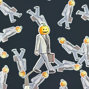 Puede incluir: Pegatinas de Halloween que representan a una persona con cabeza de calabaza con traje gris y maletín. Las pegatinas están sobre un fondo oscuro, una de ellas sujeta con pinzas.