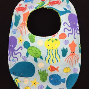 May include: A light blue baby bib with a sea life pattern. The bib features seahorses, jellyfish, starfish, sea turtles, whales, and octopuses. The bib has a green trim and a red button.