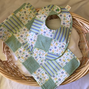 May include: A set of two baby bibs and a burp cloth in a wicker basket. The bibs and burp cloth are made of a patchwork fabric with a green, blue, and white pattern. The pattern features small yellow and white flowers and a small green and white checkered pattern.