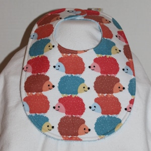 May include: A white baby bib with a pattern of colorful hedgehogs. The hedgehogs are in shades of red, orange, brown, and blue, with smiling faces. The bib has a snap closure and a curved shape to catch spills.