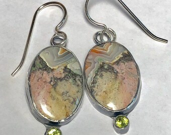 May include: A pair of silver earrings with oval-shaped agate stones set in silver. Each stone has a unique pattern of brown, white, and pink colors. A small green gemstone hangs from the bottom of each earring.